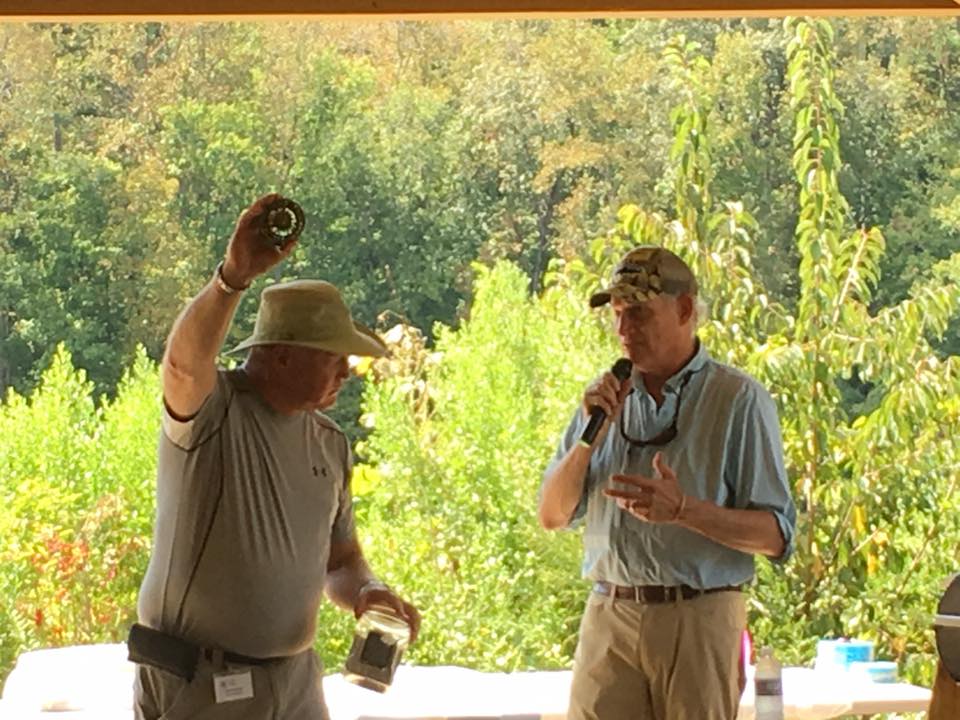 Two men are under a shelter in an outdoor setting, one holding a jar and looking at it, while the other is speaking into a microphone. The background shows dense greenery.