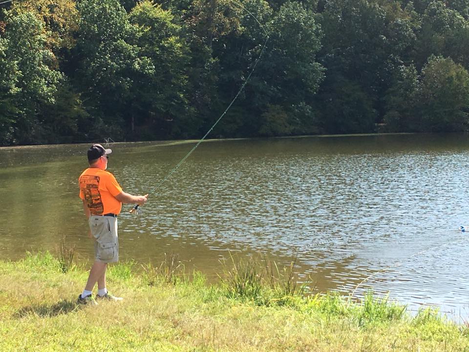 A person wearing an orange shirt and shorts is fishing at the edge of a pond surrounded by trees.
