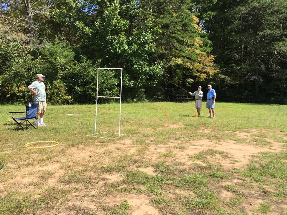 Three people in an open grassy area, involved in a fly-casting activity. One person is casting a line, another is assisting, and the third stands nearby. A target framework and hula hoops are on the ground.