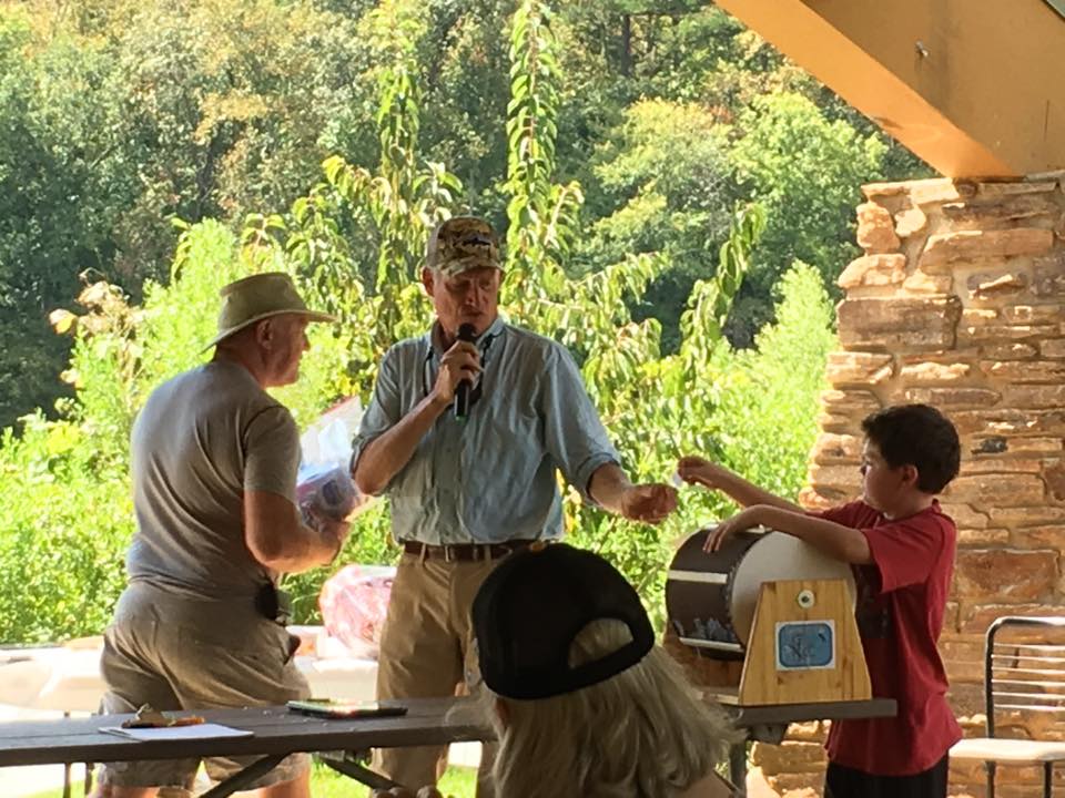 A man speaking into a microphone stands under a pavilion, while a boy and an older man operate a raffle drum in a park setting.