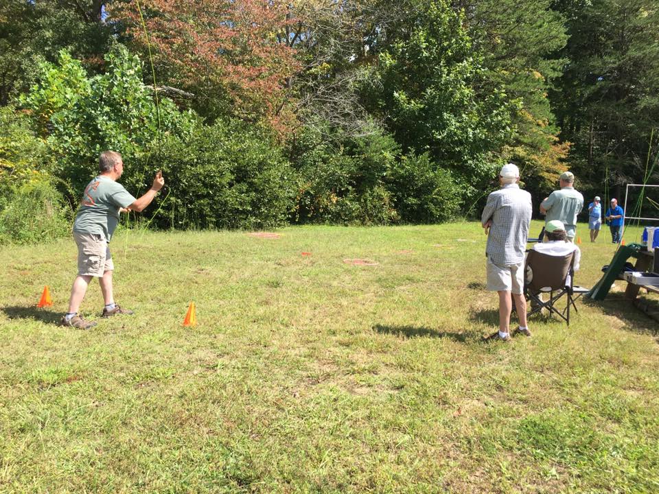 A man throws a horseshoe towards a target while a few others, one of whom is seated, watch him. The scene takes place on a grassy area with trees in the background.