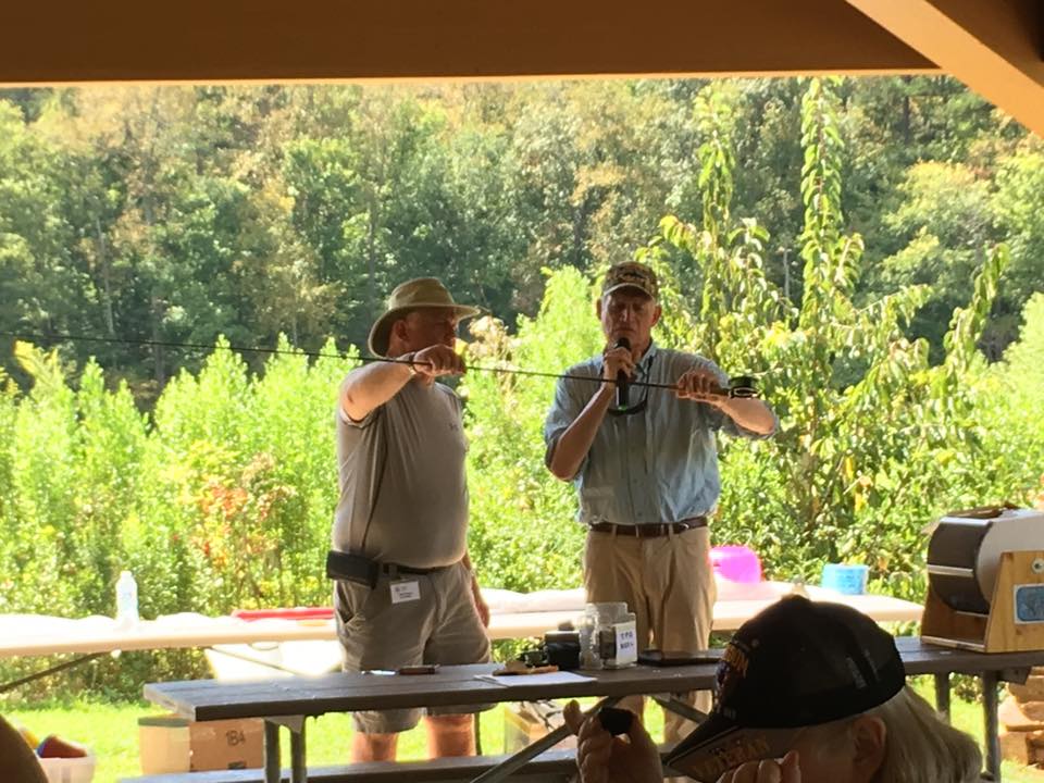 Two men are standing under a shade structure, one holding a fishing rod as the other speaks into a microphone. A green, wooded area is visible in the background.