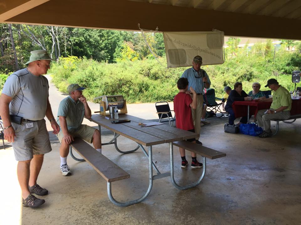 A group of people sits and stands around a picnic table under a pavilion in a park, engaging in conversation and activity.