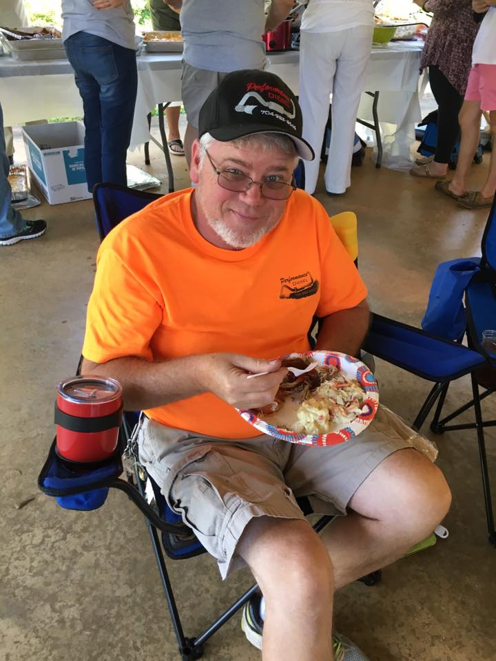 A man in a bright orange shirt and black cap sits on a folding chair, smiling while holding a plate of food. A tumbler is placed on the armrest of his chair. Other people and a buffet table are in the background.
