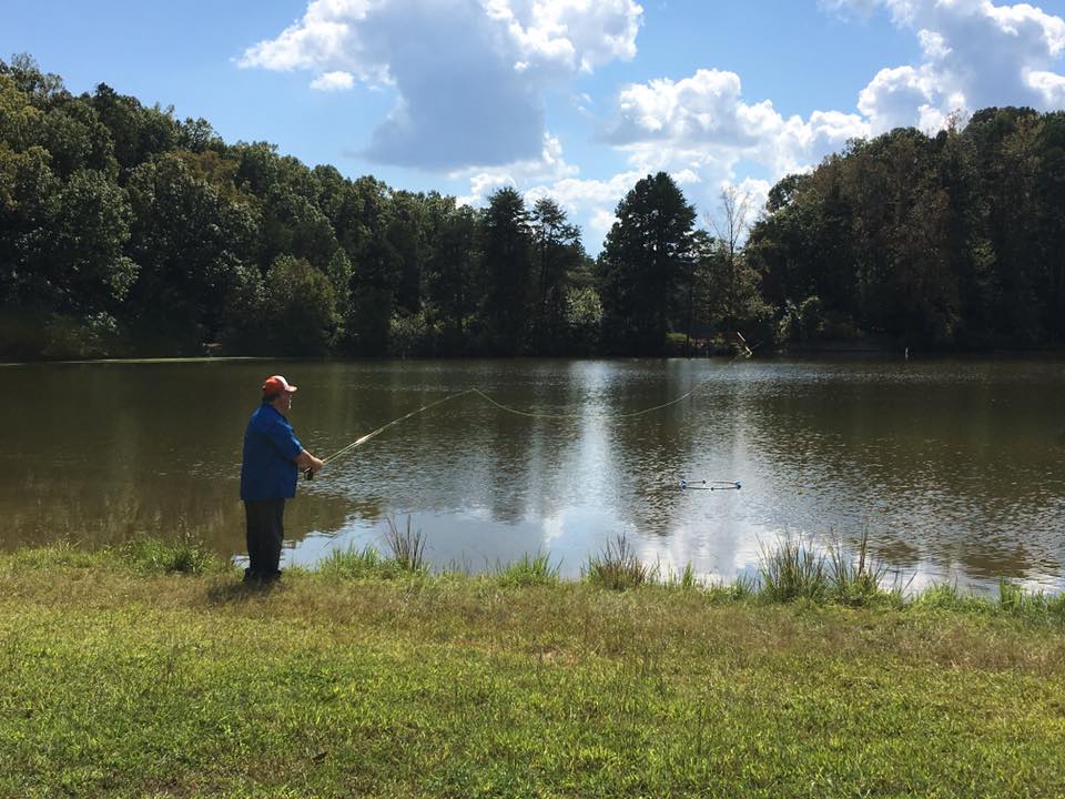 Person wearing a blue shirt and red cap is fishing on the shore of a calm lake surrounded by trees on a sunny day with scattered clouds.