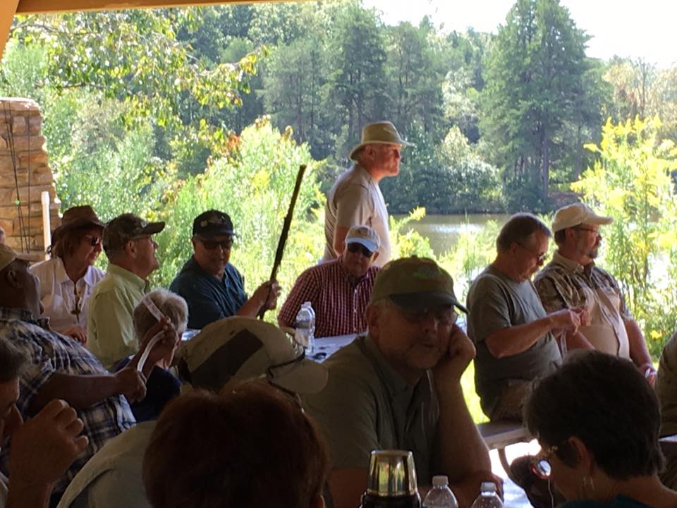 A group of people, mostly wearing hats, sit and stand under a covered outdoor area near a lake and trees on a sunny day.