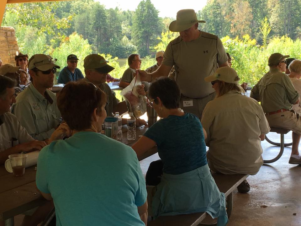 A group of people are seated at picnic tables under a pavilion in a park, engaged in conversation. Some people are standing and interacting. The background shows trees and greenery.