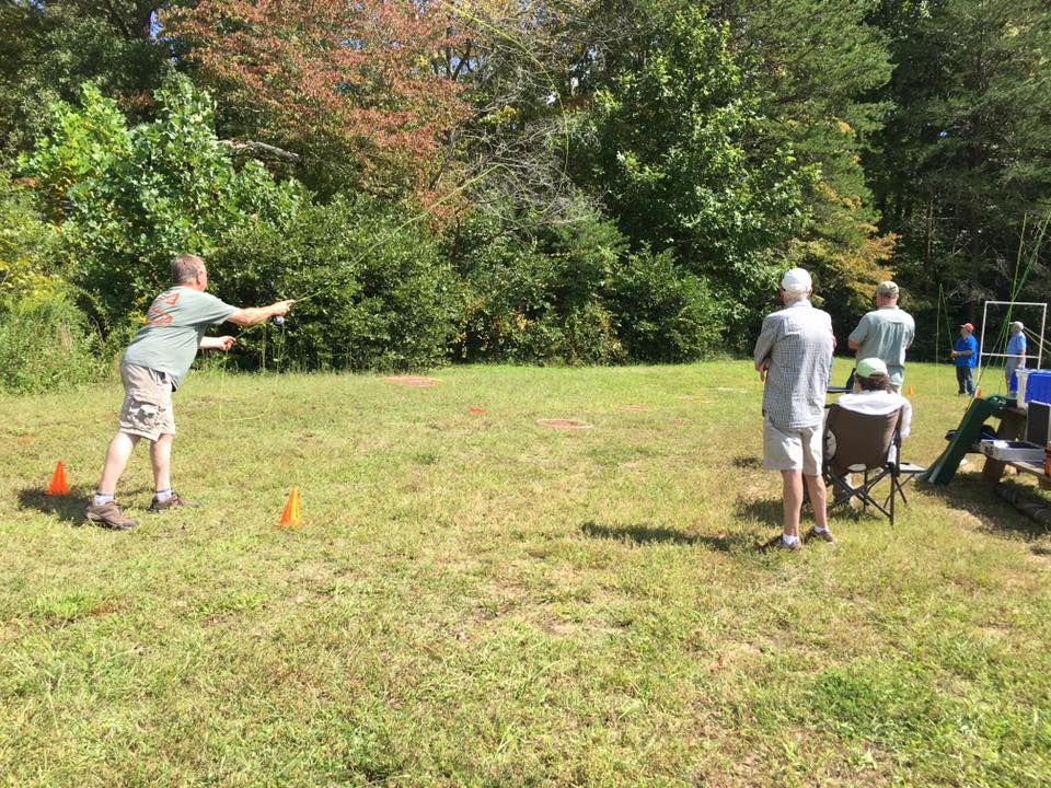 Four people are outside in a grassy area. One person is throwing an object, with three others watching. The scene is surrounded by trees and there are two orange cones on the ground.