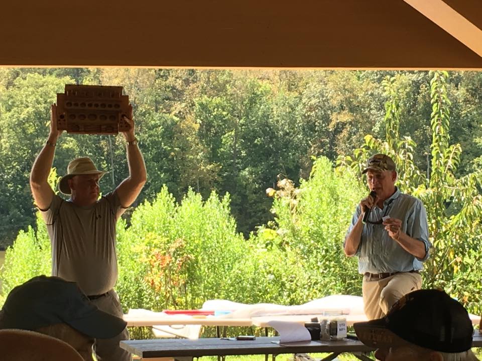 Two men stand under a canopy. One is holding up a wooden object while the other speaks into a microphone. Trees and shrubs are visible in the background.