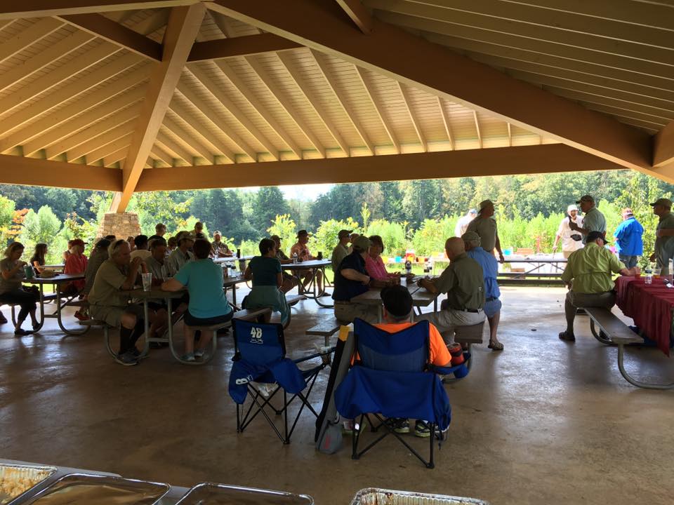 A group of people gather under a large, open-sided pavilion in a park, sitting at picnic tables and chatting. Trees and greenery are visible in the background.
