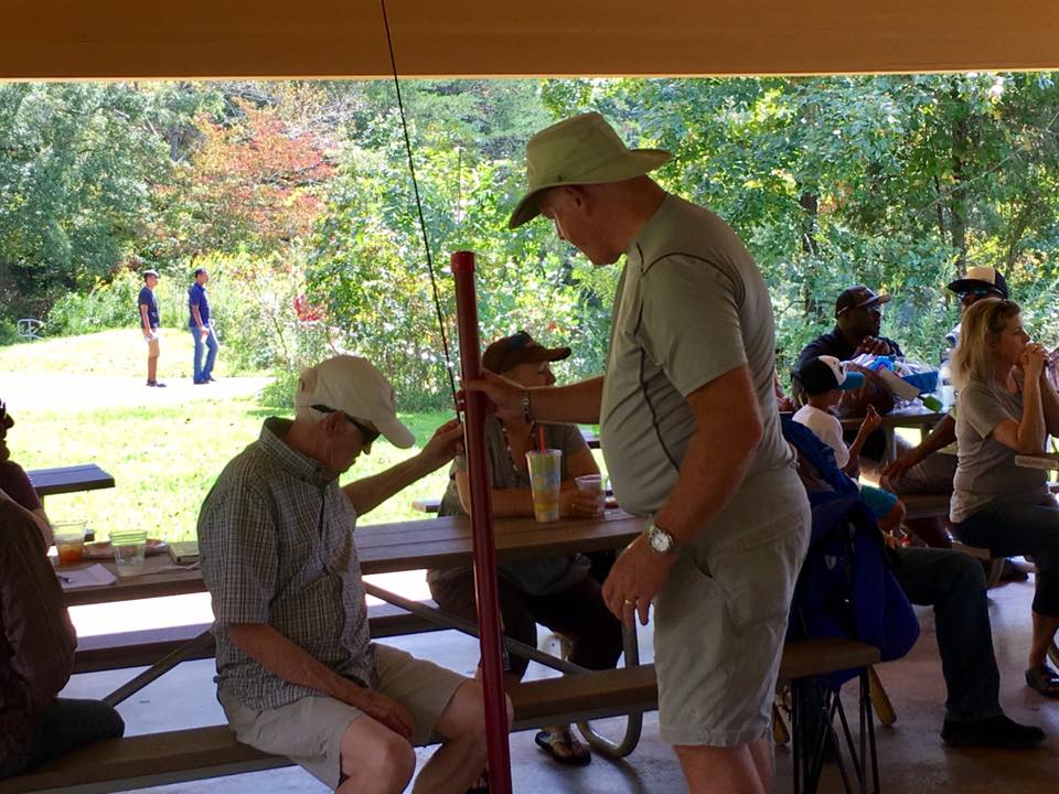 A group of people are seated at picnic tables under a shelter in a park. Two men in hats interact near a red post, while others sit, eat, and converse. Trees and greenery are in the background.