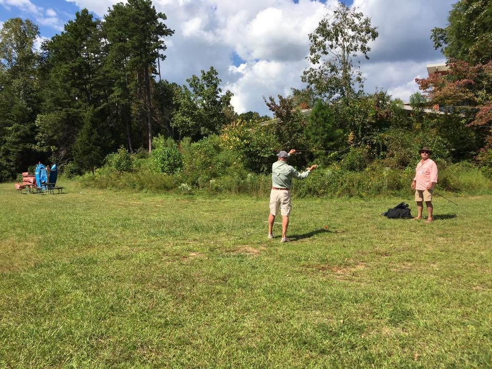 Two people are fly fishing in an open grassy area surrounded by trees on a sunny day. There are three blue folding chairs and a table in the background.