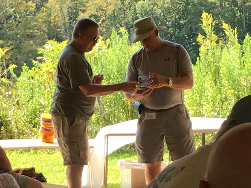 Two men are standing outdoors near a table and examining a small object together. They are dressed in casual clothes and are surrounded by greenery.
