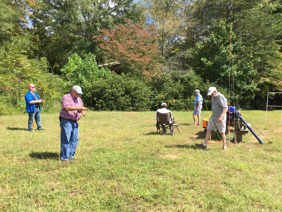 Several people are spread out in a grassy area, engaged in various activities such as sitting on chairs and looking at their devices, with trees and a clear sky in the background.