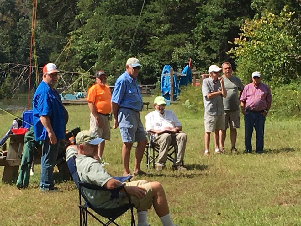 Several men standing and seated outdoors near a body of water, with trees and fishing gear in the background. One man is seated in a folding chair while others stand in a line.