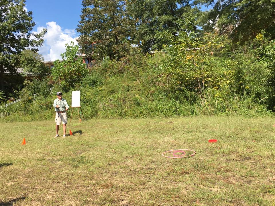 A person stands on a grassy field near two orange cones, holding a frisbee. In the distance, there's a whiteboard on a stand and lush green trees. A hula hoop and red bucket are on the ground.