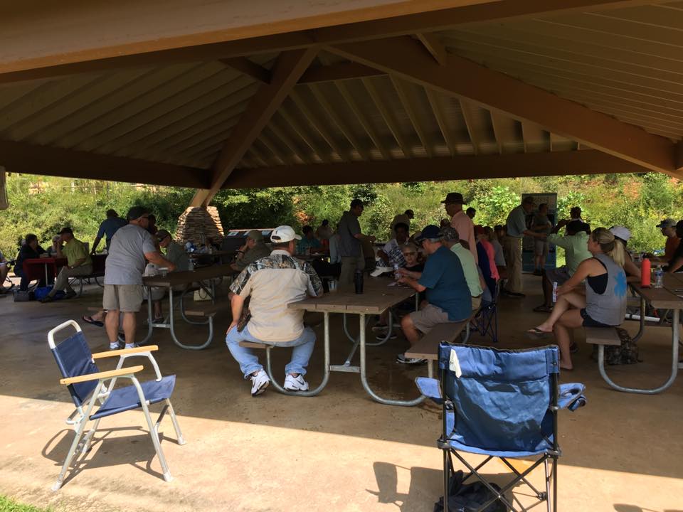 A group of people gathered under a pavilion in a park, sitting at picnic tables and chatting. Some are standing while others are seated, with various chairs and items on the tables.