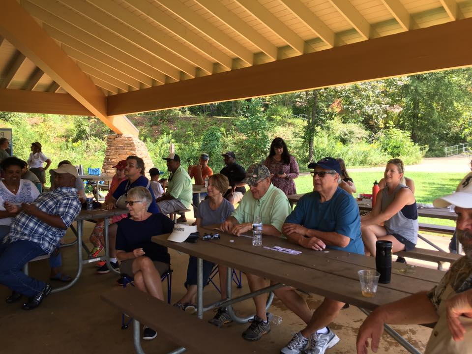 A group of people are seated at picnic tables under a pavilion, attentively watching something off-camera. They are outdoors in a wooded area, and the weather appears to be clear and sunny.