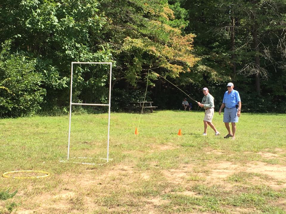 Two people are outdoors practicing fly fishing casting. One is holding a fishing rod and casting, while the other watches. There is a white frame and cones set up on the grass, surrounded by trees.